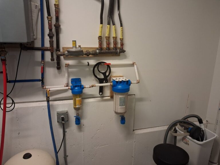 Two whole-house filter housings with blue caps plumbed on a utility room wall